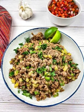 A plate of seasoned ground meat garnished with fresh herbs, accompanied by lime slices, garlic, and a bowl of diced vegetables.