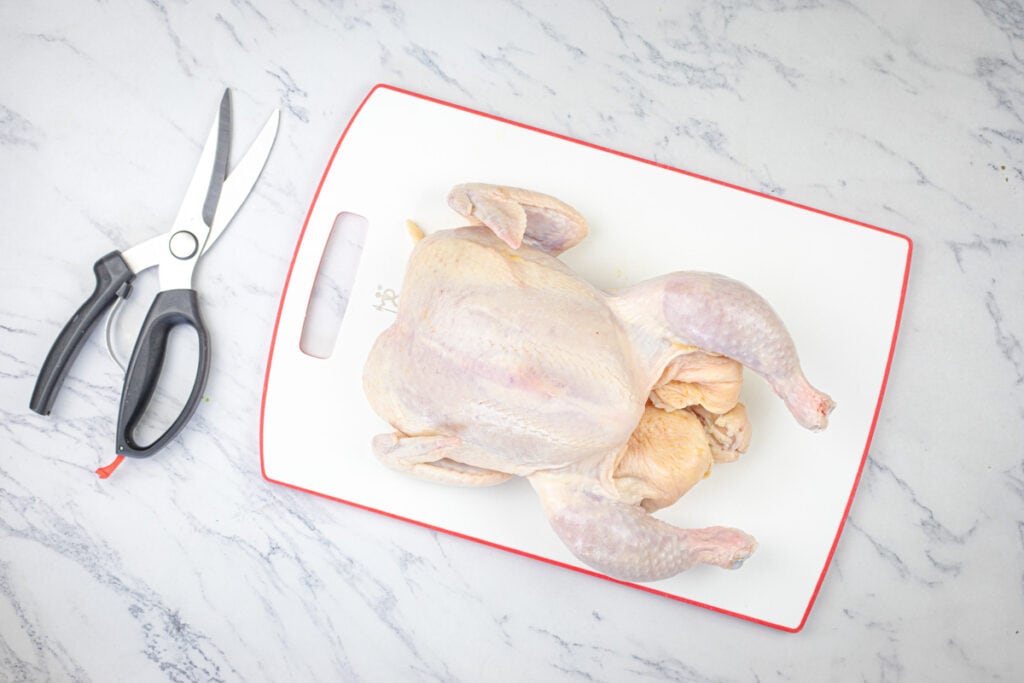 Raw whole chicken on a white cutting board next to kitchen shears and a knife.