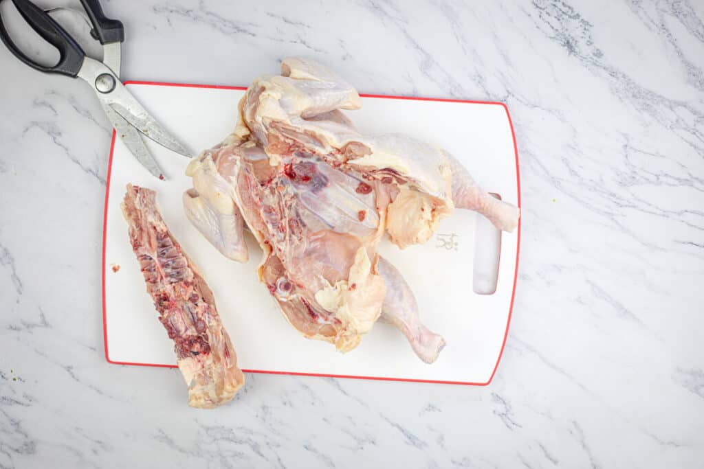 Raw chicken being prepared on a cutting board with kitchen scissors and a bone to the side.