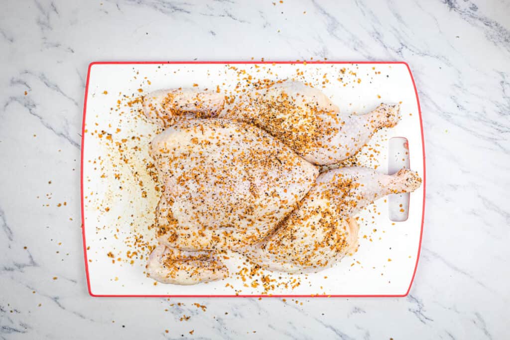 A raw chicken spatchcocked and seasoned on a cutting board.