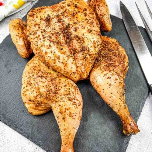 Roasted chicken with seasoning displayed on a slate board with cutlery on the side.