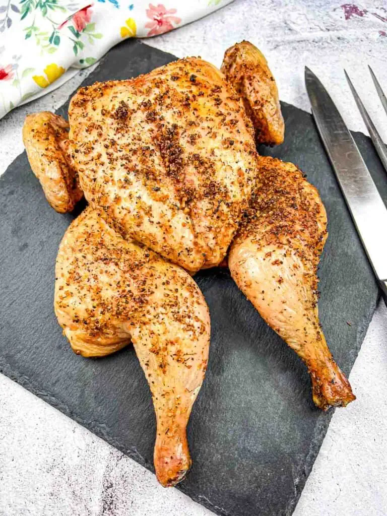 Roasted chicken with seasoning displayed on a slate board with cutlery on the side.