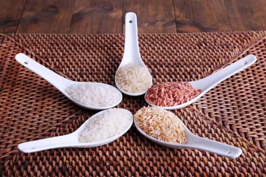Types of rice displayed on white spoons over a woven mat.