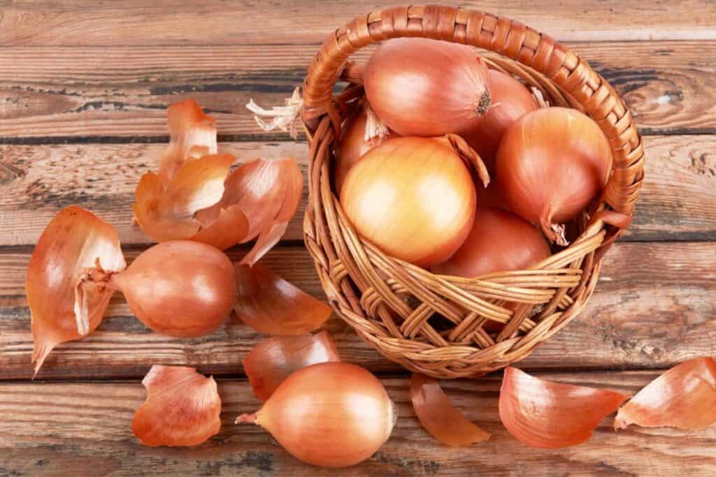 A basket of whole onions with some scattered onion skins on a wooden surface.