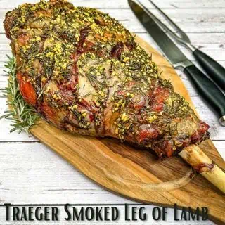 Herb-crusted Traeger Smoked Leg of Lamb on a wooden cutting board with kitchen knives.