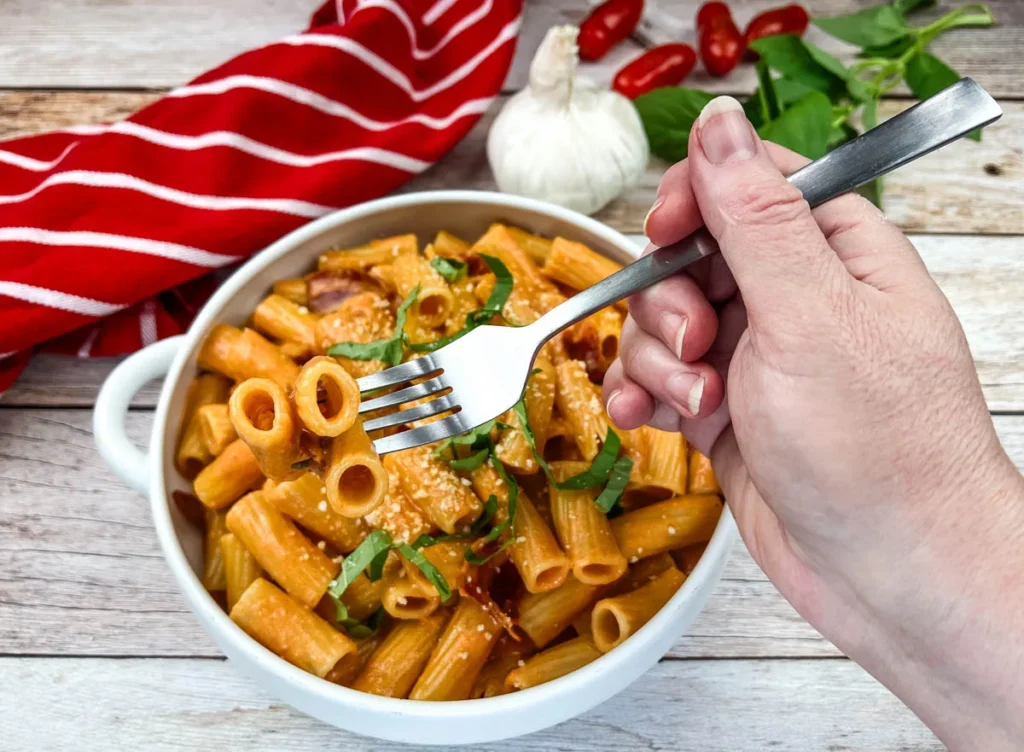 A hand holding a fork with pasta in a white bowl, surrounded by garlic, chili peppers, and a red-striped napkin on a wooden surface.