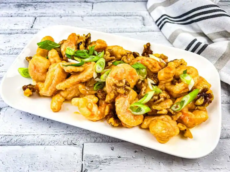 A plate of crispy orange chicken garnished with green onions and walnuts, served on a white rectangular dish on a rustic table.