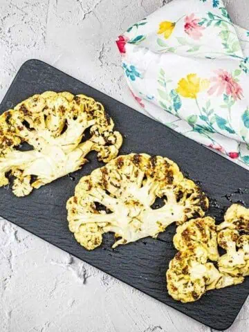 Roasted cauliflower steaks on a black serving slate with a floral napkin on the side.