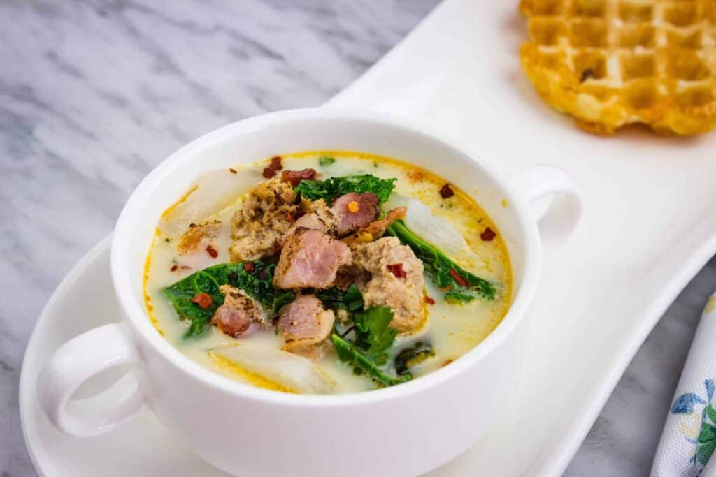 A bowl of creamy soup with chunks of sausage, bacon, and kale, served beside a piece of waffle on a white rectangular plate.