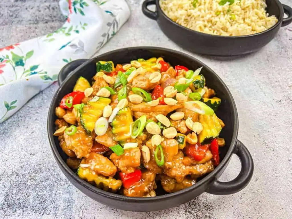 A bowl of kung pao chicken topped with scallions and peanuts, served beside a bowl of rice on a textured surface.