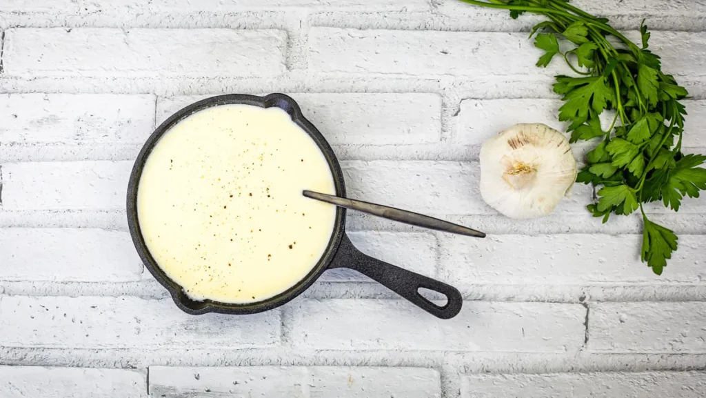 A skillet of creamy cheese fondue with a spoon, paired with fresh garlic and parsley, set against a white brick backdrop.