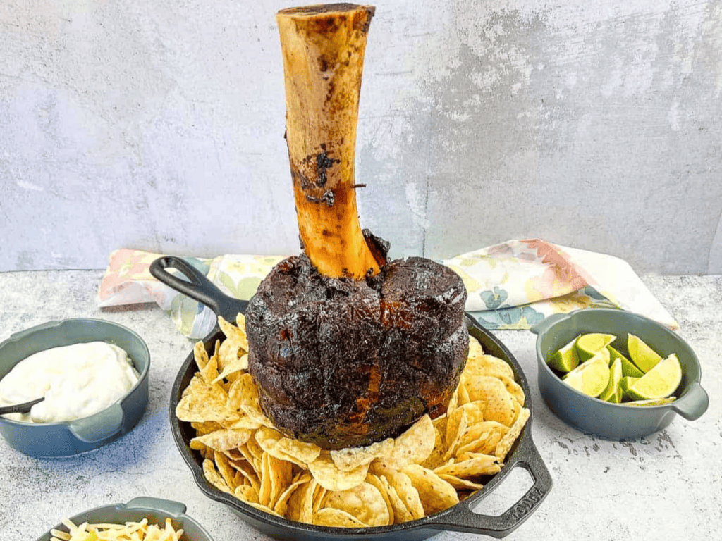 A large roasted meat shank on a bone, displayed upright in a skillet surrounded by nachos, with bowls of lime wedges and sauce nearby.