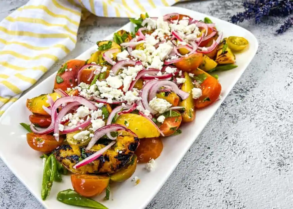 A colorful grilled vegetable salad with cherry tomatoes, onions, zucchini, and crumbled feta cheese on a white plate.