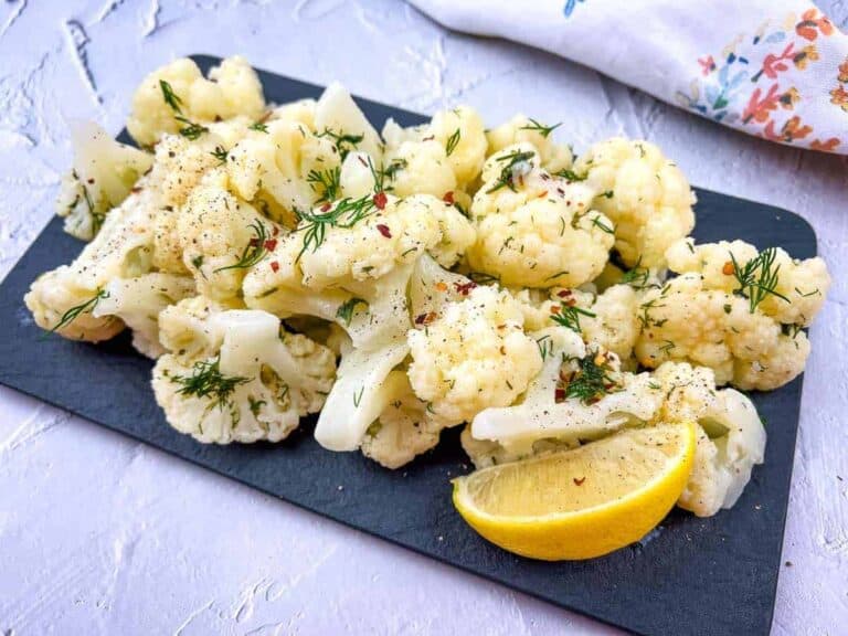 A plate of steamed cauliflower garnished with dill and lemon wedges on a slate board.