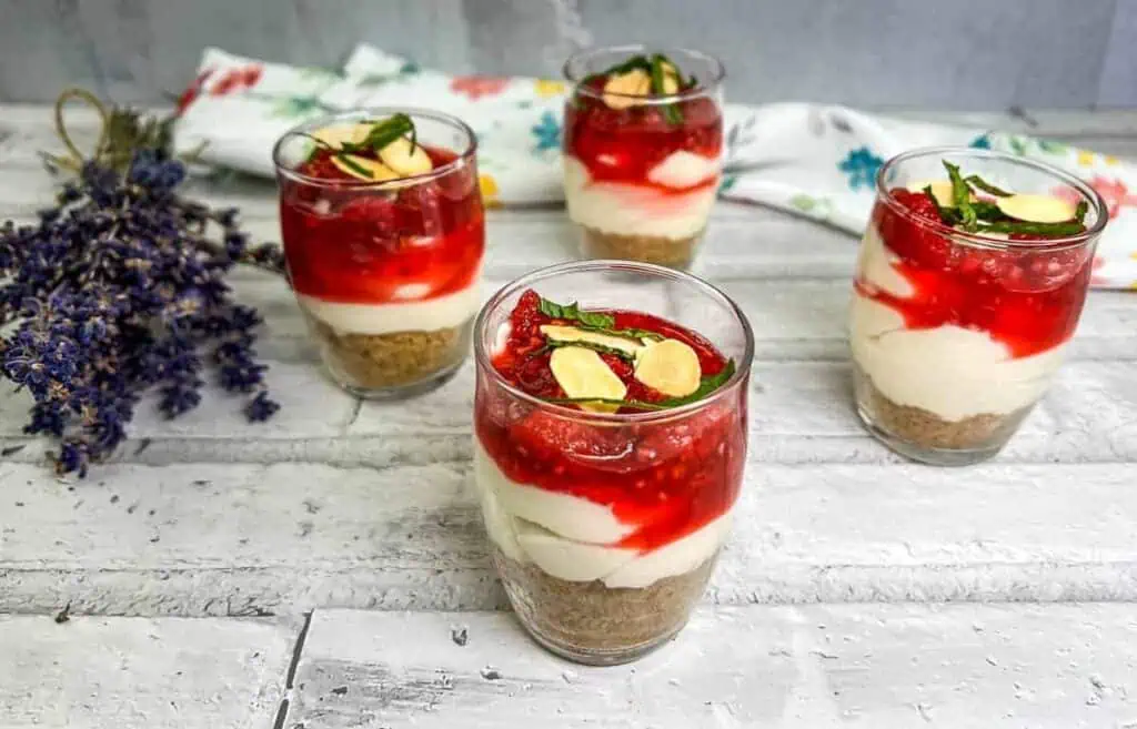 Four glasses of layered dessert with a biscuit base, white cream, and red fruit topping, garnished with mint leaves, on a rustic table with lavender in the background.