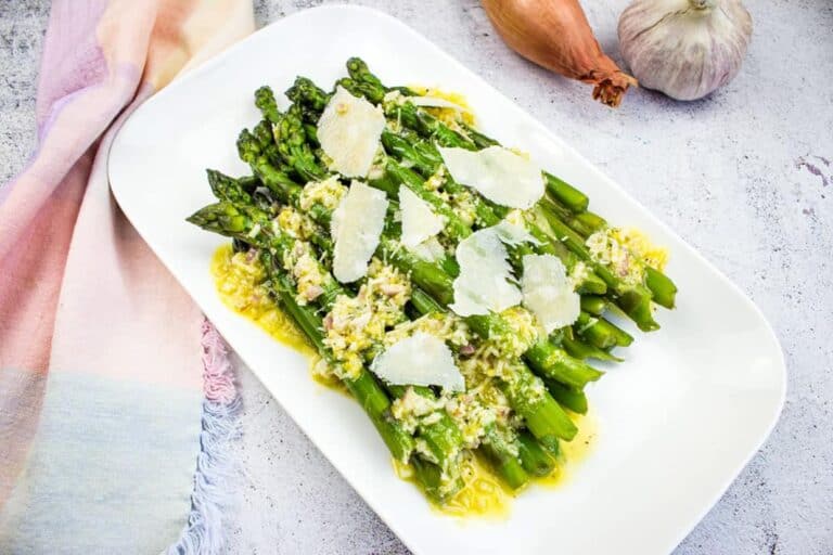 A dish of cooked asparagus topped with shaved parmesan and a light sauce, served on a white rectangular plate with garlic and an onion in the background.