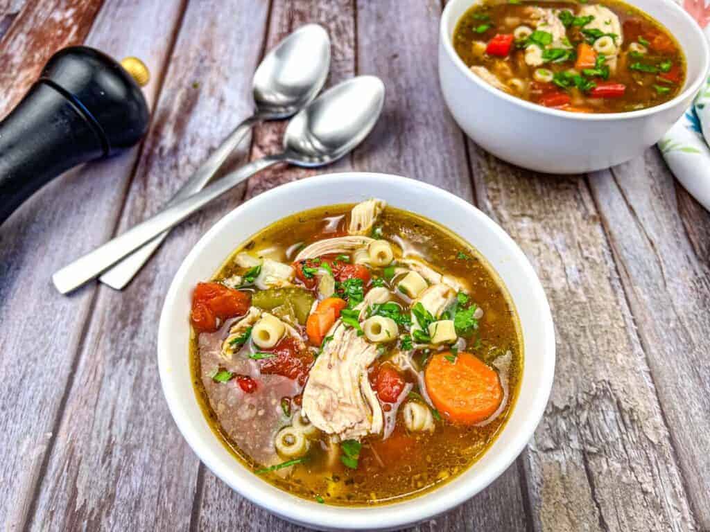 Two bowls of Copycat Carrabba's Mama Mandola's Sicilian Chicken Soup, accompanied by two spoons, a black pepper grinder, and a napkin.