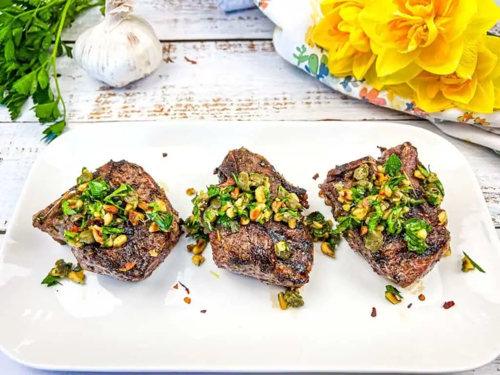 Three grilled lamb chops topped with garlic and herb seasoning on a white plate, accompanied by yellow flowers and garlic cloves on a wooden table.