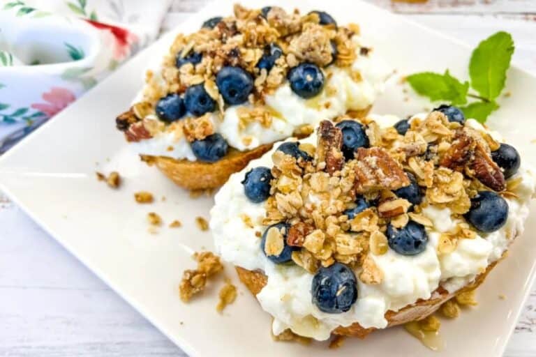 A close-up of two pastries topped with a generous amount of cottage cheese, fresh blueberries, granola, and a drizzle of honey. A fresh green mint leaf decorates the plate.