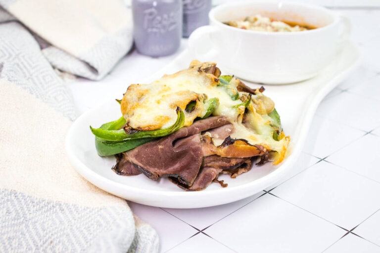 A plate of roast beef topped with melted cheese and green peppers, next to a bowl of soup on a white, tiled surface.