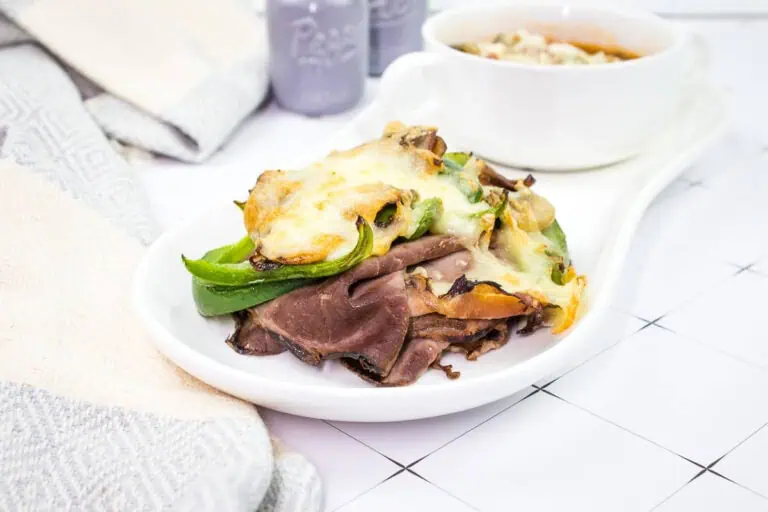A plate of roast beef topped with melted cheese and green peppers, next to a bowl of soup on a white, tiled surface.