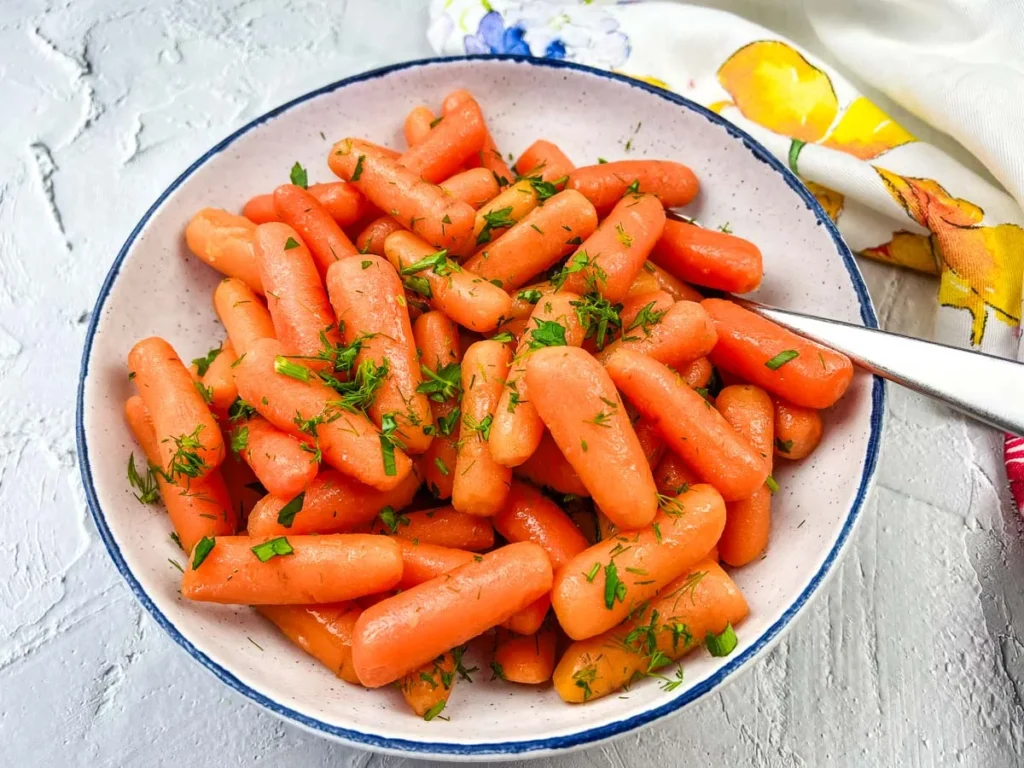 Bowl of glazed baby carrots sprinkled with fresh dill, served on a white and blue rimmed plate with a spoon on the side.