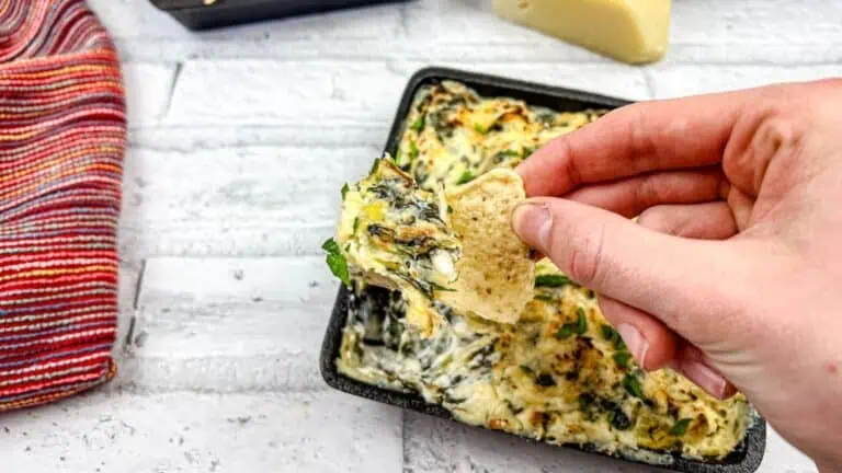 A hand holding a pita chip dipped in a cheesy spinach artichoke dip, with the dip dish and a striped cloth in the background.