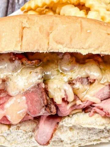 A roast beef sandwich with melted cheese on a hoagie roll, accompanied by crinkle-cut fries and a bowl of brown dipping sauce on the side.
