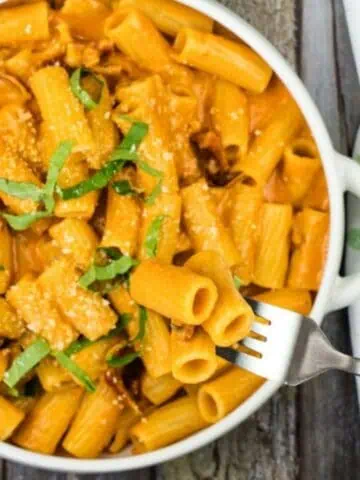 A white bowl of rigatoni pasta with creamy sauce, garnished with cheese and green herbs, is placed on a wooden surface. A hand is holding a fork with a piece of pasta.