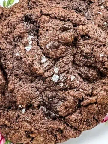 A close-up of a large chocolate cookie sprinkled with coarse salt, placed on a decorative plate with pink rose patterns.