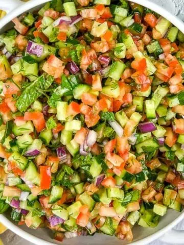 A bowl of chopped vegetable salad with cucumbers, tomatoes, red onions, and parsley, next to a yellow checkered cloth and fresh parsley.