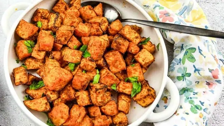 A white bowl filled with seasoned roasted sweet potato cubes, garnished with chopped herbs, with a metal serving spoon on the side and a printed napkin partially visible.