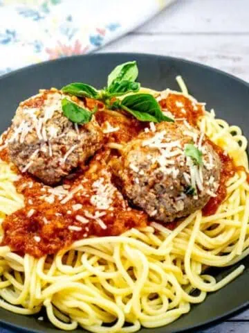 A plate of spaghetti topped with two meatballs, marinara sauce, grated cheese, and basil garnish.