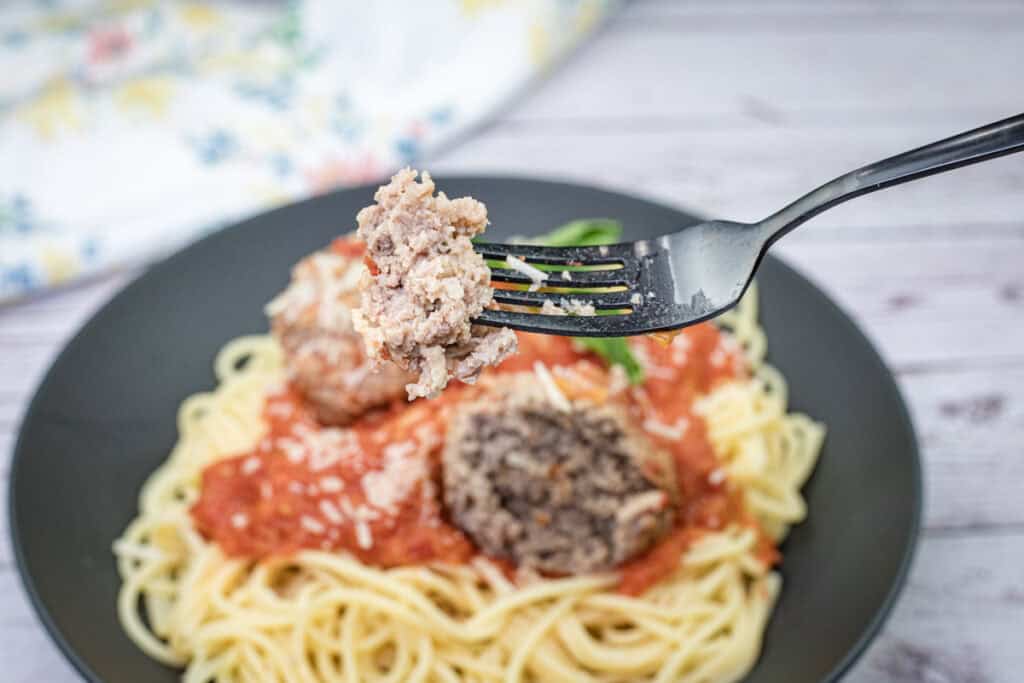 A fork holds a giant meatball above a plate of spaghetti topped with tomato sauce and shredded cheese.