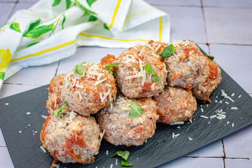 A stack of giant, seasoned meatballs garnished with grated cheese and fresh herbs is served on a black rectangular slate plate. A cloth with a lemon pattern is visible in the background.
