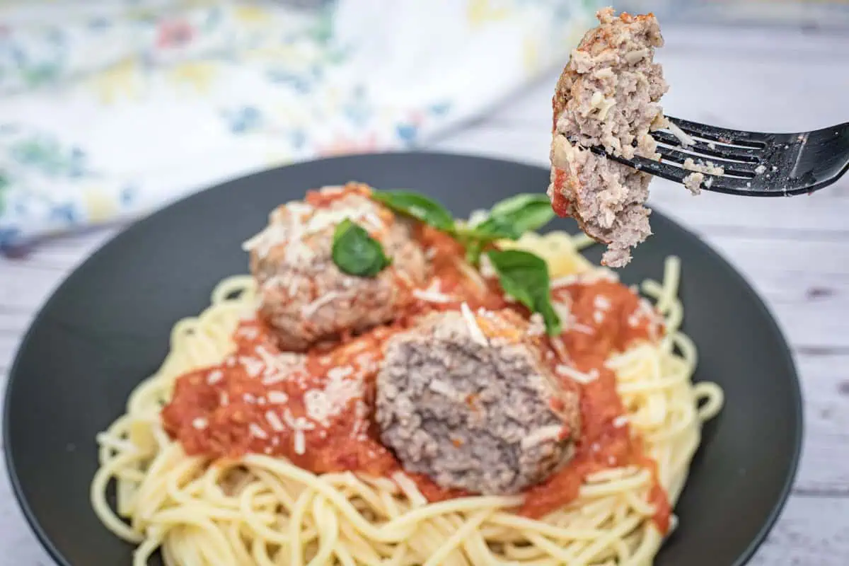 A fork holding a cut piece of giant meatball over a plate of spaghetti topped with massive meatballs, marinara sauce, and garnished with grated cheese and basil leaves.