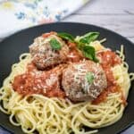 A plate of spaghetti topped with tomato sauce, two large meatballs, grated cheese, and a sprig of basil.