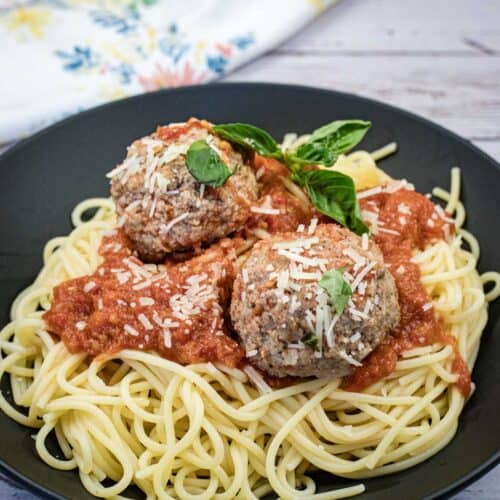 A plate of spaghetti topped with tomato sauce, two large meatballs, grated cheese, and a sprig of basil.