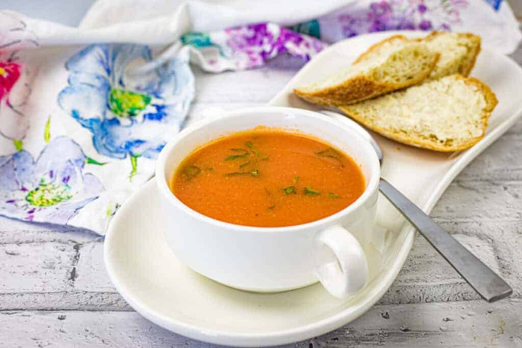 A bowl of old-fashioned tomato soup garnished with herbs, served with slices of crusty bread on a white plate.