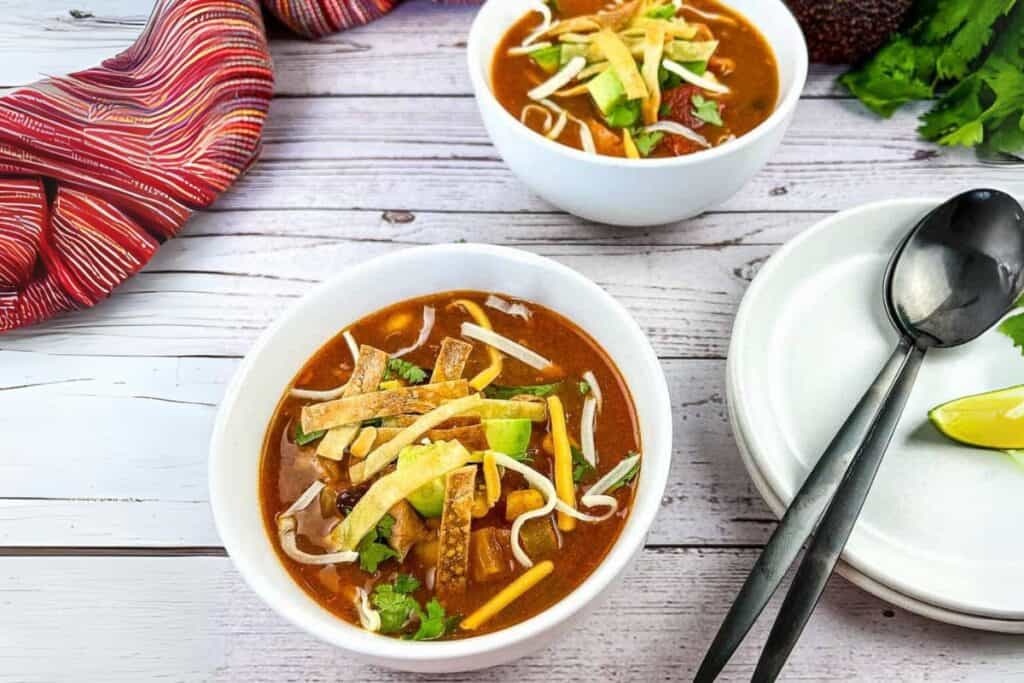 Two bowls of Turkey Tortilla Soup with a plate and spoons beside on a wooden table.