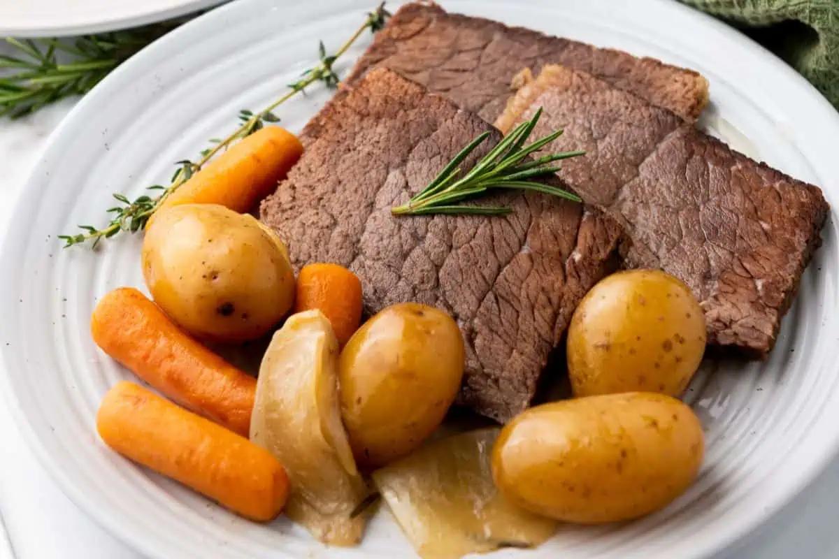 A plate of slow cooker rump roast with potatoes, carrots, onions, and a sprig of rosemary on a white dish.