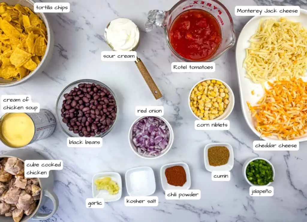 Ingredients for Cheesy Mexican Casserole laid out on a marble surface, including tortilla chips, sour cream, Rotel tomatoes, cheeses, chicken, black beans, red onion, corn, garlic, seasonings, and jalapeño.