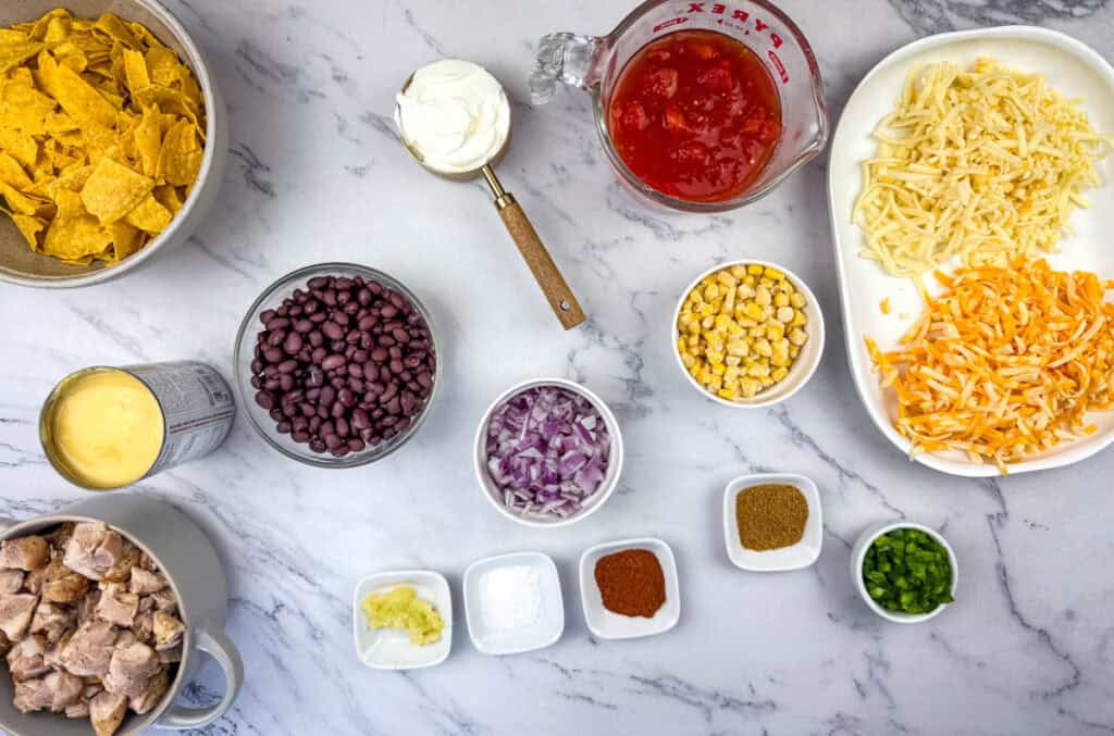 Ingredients for Cheesy Mexican Casserole, including tortilla chips, sour cream, canned beans, corn, cheese, diced tomatoes, chicken pieces, chopped onions, and seasonings like cumin and chili powder on a counter.