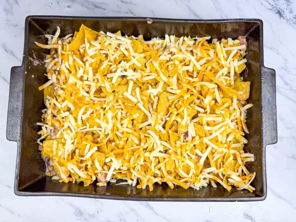 A baking dish filled with tortilla chips, covered with shredded cheddar and mozzarella cheese, on a marble countertop.