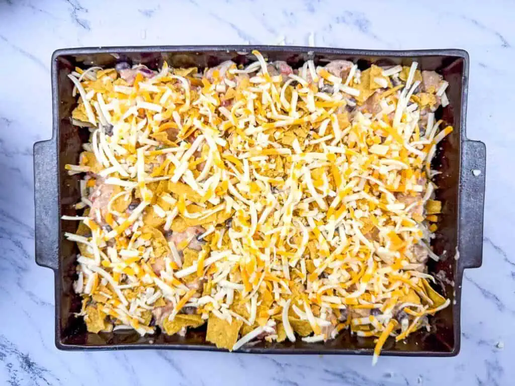 Baking dish with uncooked casserole of shredded cheese, crumbled tortilla chips, beans, and other ingredients on a marble countertop.