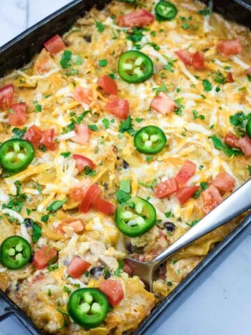 A Cheesy Mexican Casserole topped with melted cheese, jalapeño slices, diced tomatoes, and chopped herbs, with a serving spoon placed on the side.
