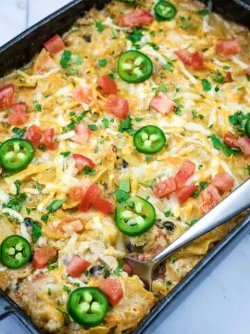 A Cheesy Mexican Casserole topped with melted cheese, jalapeño slices, diced tomatoes, and chopped herbs, with a serving spoon placed on the side.