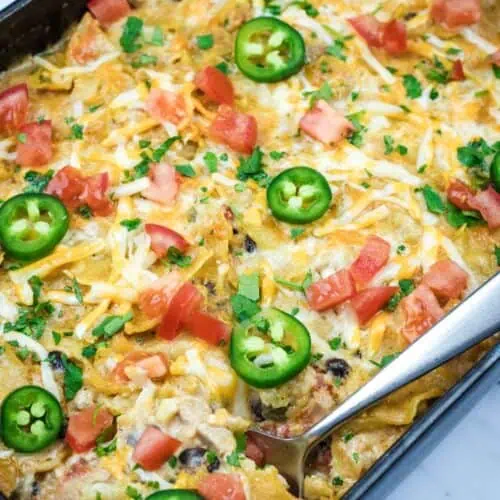 A Cheesy Mexican Casserole topped with melted cheese, jalapeño slices, diced tomatoes, and chopped herbs, with a serving spoon placed on the side.