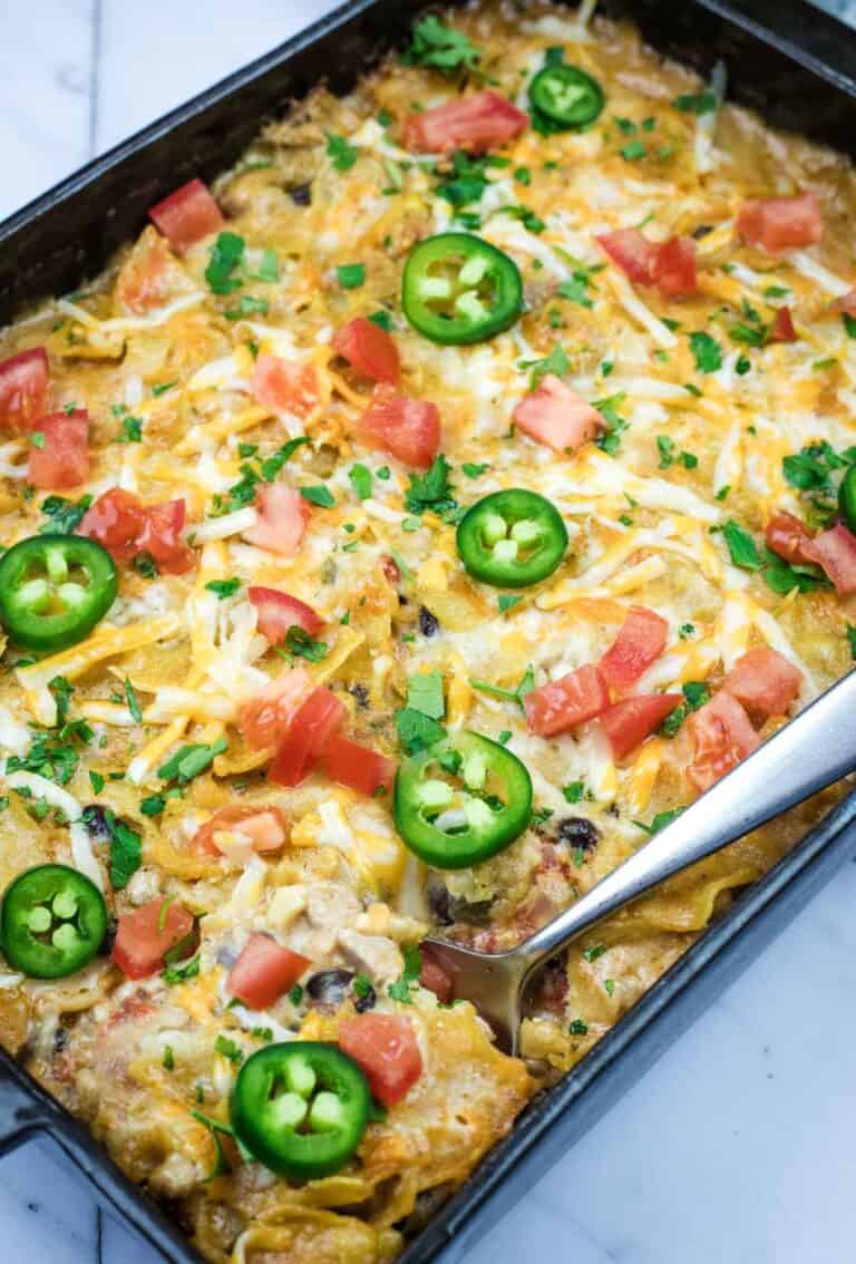 A Cheesy Mexican Casserole topped with melted cheese, jalapeño slices, diced tomatoes, and chopped herbs, with a serving spoon placed on the side.