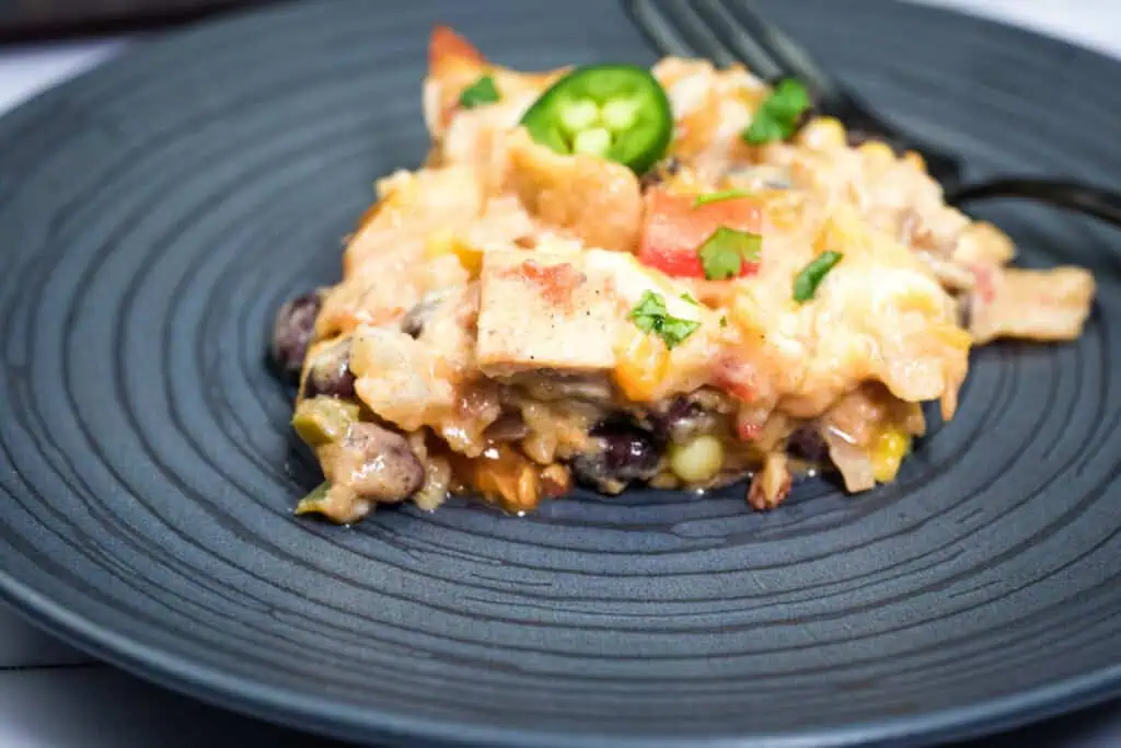 A serving of Cheesy Mexican casserole with melted cheese, vegetables, and a slice of jalapeño on a dark plate, with a fork in the background.